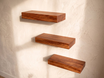 Three floating wooden shelves in a warm medium-brown finish, mounted on a textured off-white plaster wall with soft sunlight casting shadows.