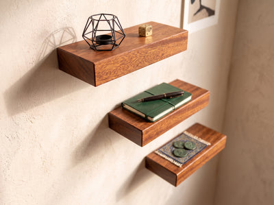 Three floating wooden shelves mounted on a textured beige wall, styled with a black geometric candle holder, a small gold cube, a green notebook with a pen, and a decorative tray with coins.