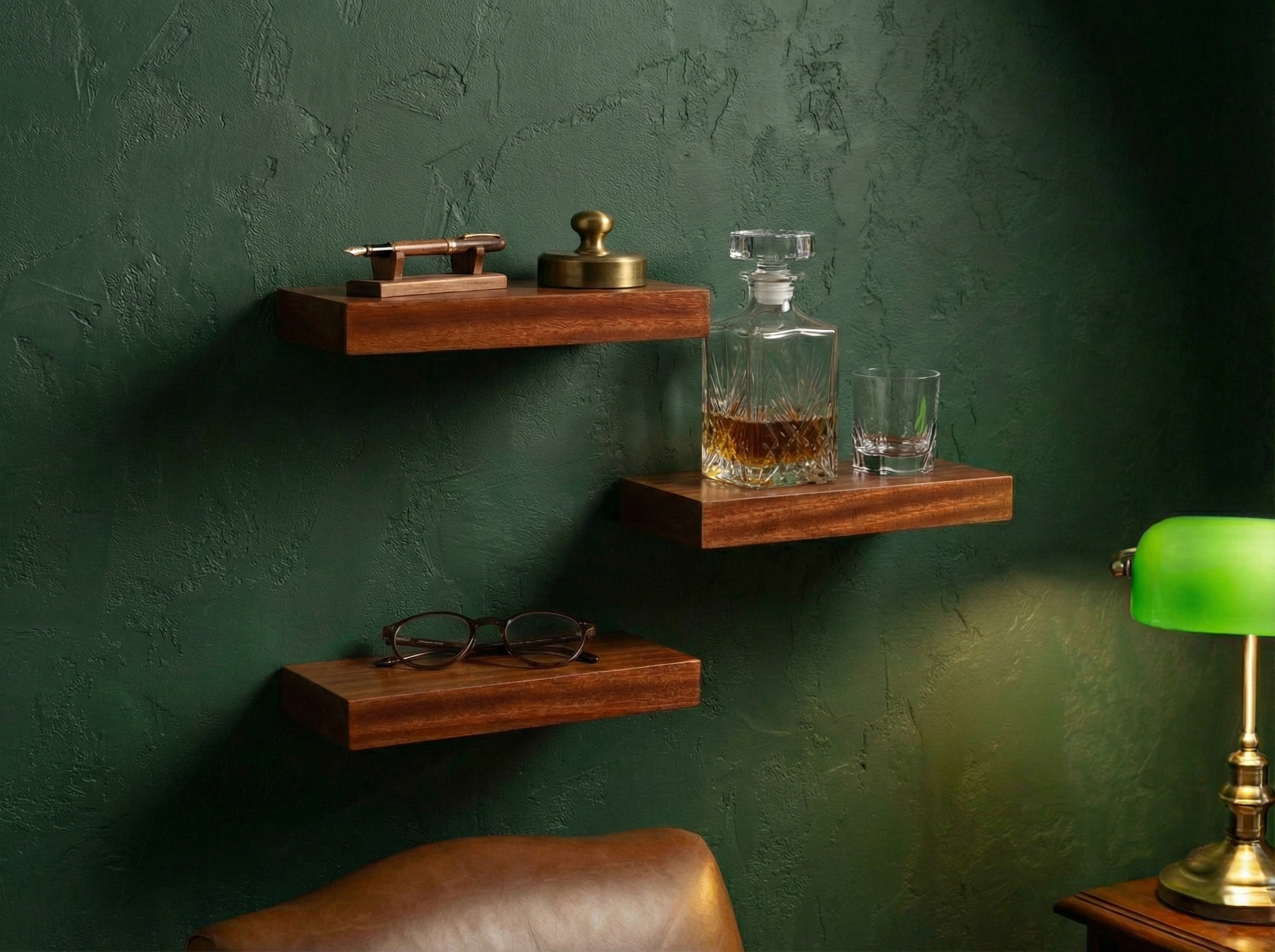 Three dark brown floating wooden shelves mounted on a textured dark green wall, styled with a brass object, a glass decanter and two glasses, and a pair of eyeglasses. A green lamp and a brown leather chair are partially visible in the foreground.