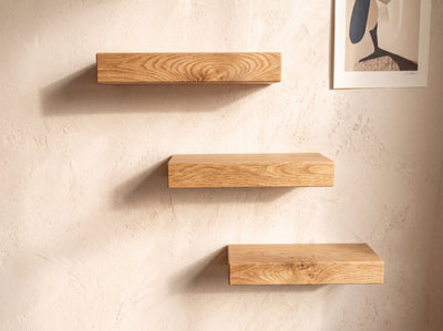 Three floating wooden shelves in varying heights mounted on a textured beige plaster wall, with a framed abstract print partially visible in the top right corner.
