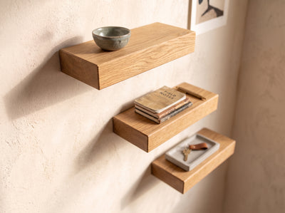 Three floating wooden wall shelves mounted on a textured beige plaster wall, styled with a small ceramic bowl, a stack of books, and a small tray with keys.